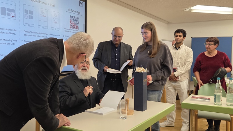 Bischof sitzt am Tisch und signiert seine neue Broschüre. Menschen stehen neben ihm in der Schlange.