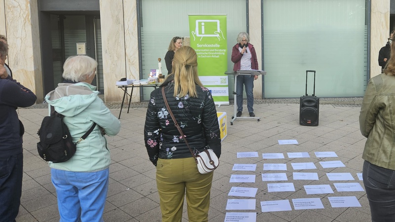 Öffentliche Veranstaltung: Eine Sprecherin steht am Pult vor Zuschauern. Auf dem Boden liegen Zettel mit Grundrechten, dahinter ein Grünes Banner.