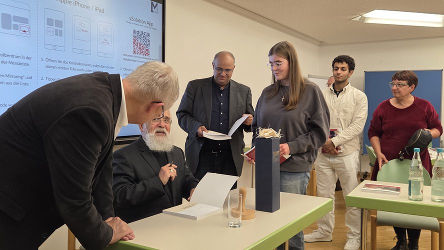 Bischof sitzt am Tisch und signiert seine neue Broschüre. Menschen stehen neben ihm in der Schlange.