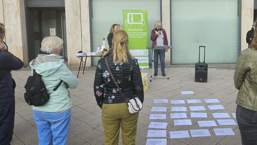 Öffentliche Veranstaltung: Eine Sprecherin steht am Pult vor Zuschauern. Auf dem Boden liegen Zettel mit Grundrechten, dahinter ein Grünes Banner.