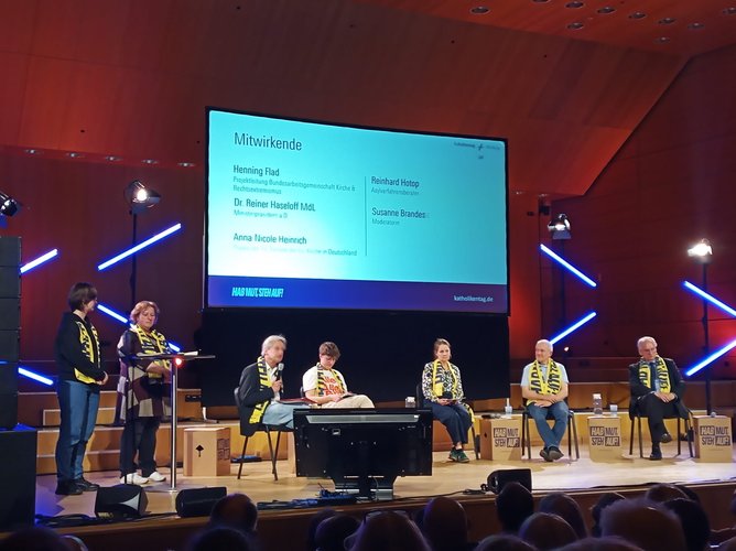 Menschen auf einem Podium, hinter ihnen ein Bildschirm mit der Beschreibung der Veranstaltung